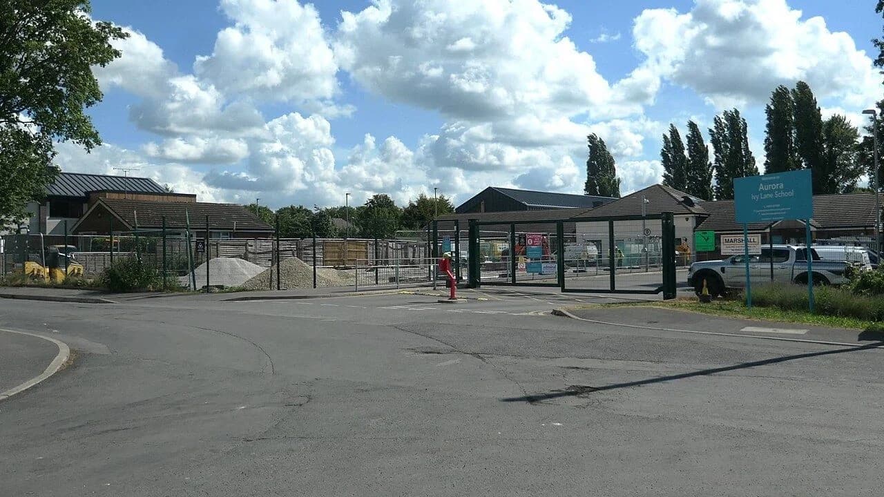A school construction site behind metal fencing, with earthworks, machinery and site buildings under a partly cloudy sky