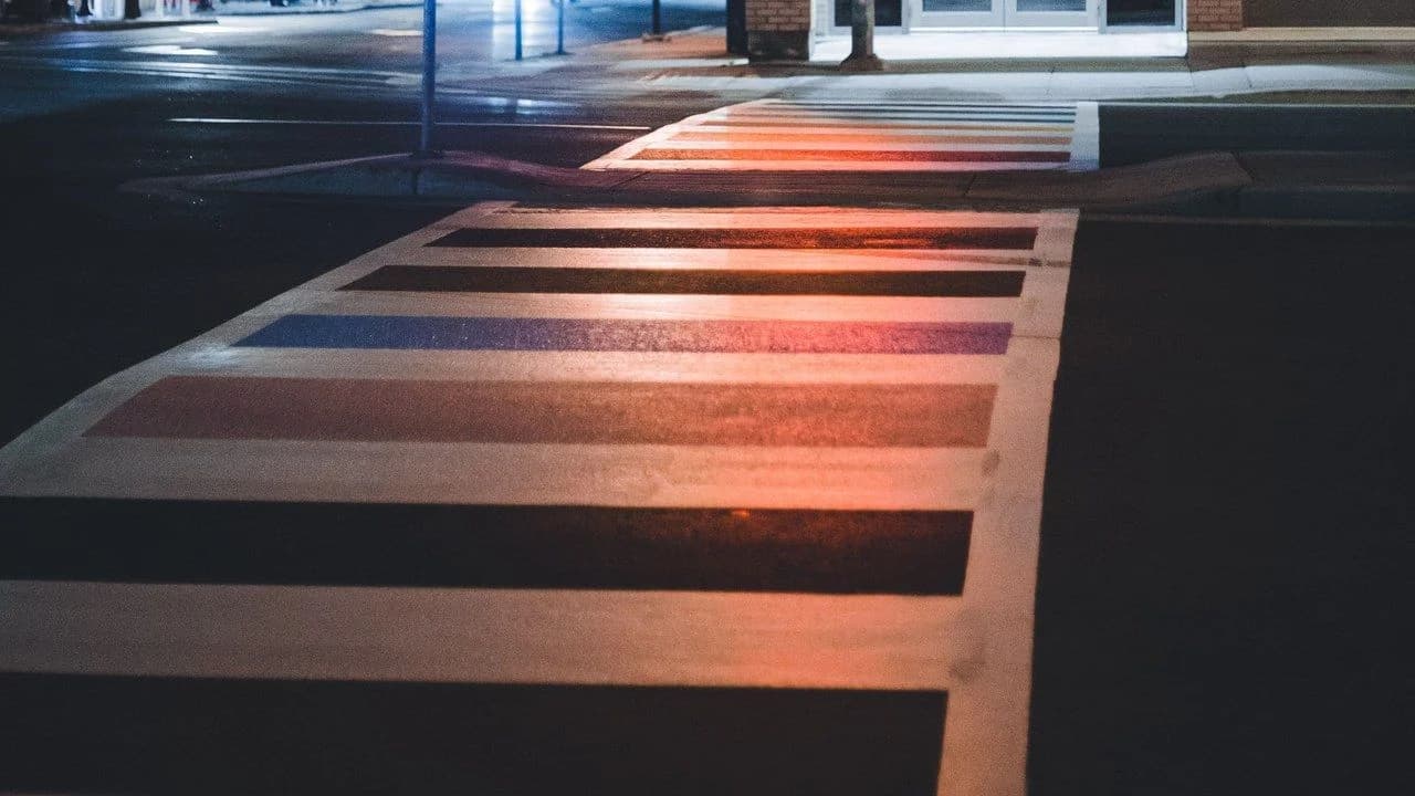 Empty night-time city intersection: dark road with lit crosswalks under the red glow of a traffic light; no people, no vehicles.
