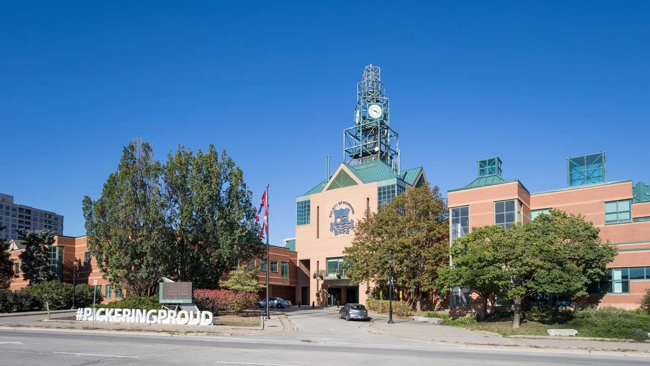 Exterior of Pickering City Hall (Pickering Civic Complex) in Pickering, Ontario — a brick civic building with a clock tower, City of Pickering crest and #PICKERINGPROUD sign under a clear blue sky; no people.